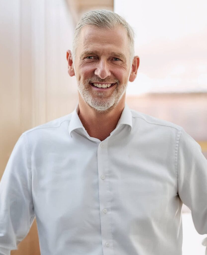 Lächelnder Mann mit grauen Haaren und weißem Hemd schaut freundlich in die Kamera.