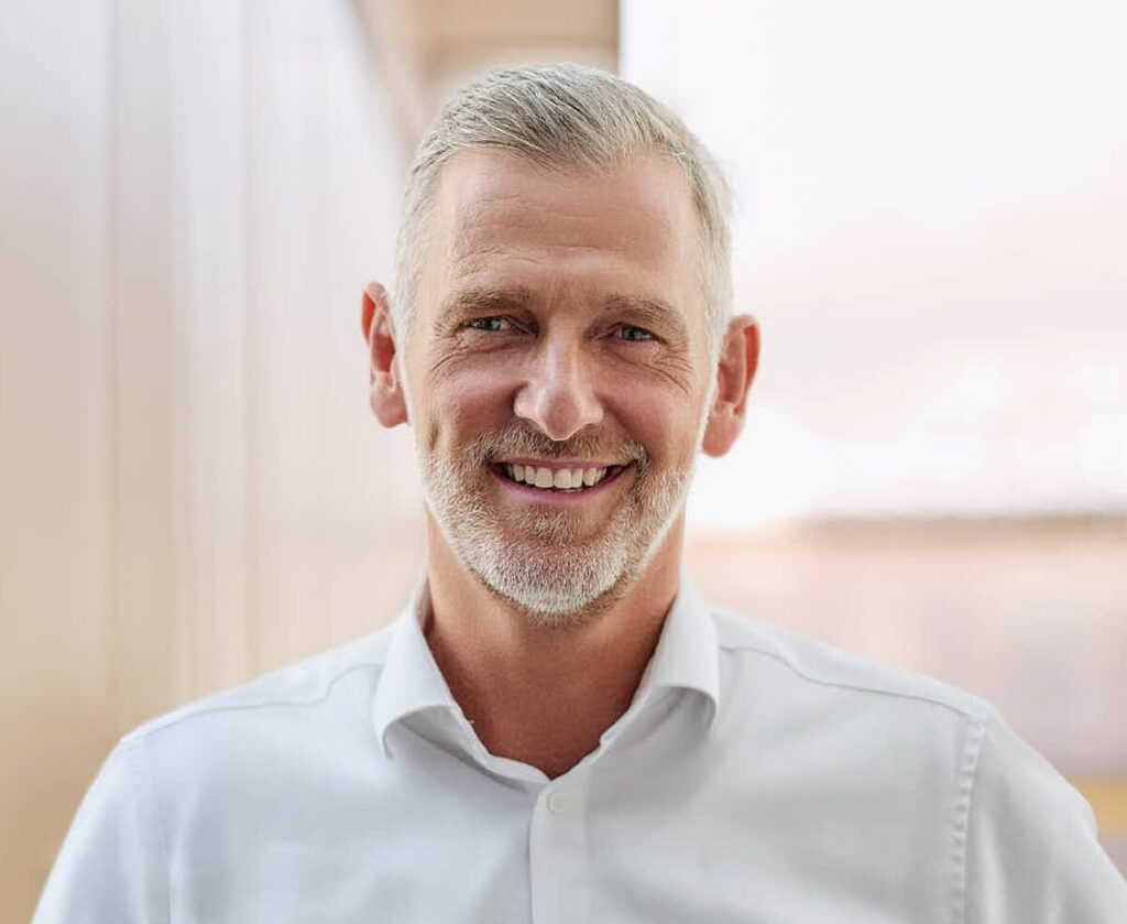 Lächelnder Mann mit grauen Haaren und Bart in einem weißen Hemd.