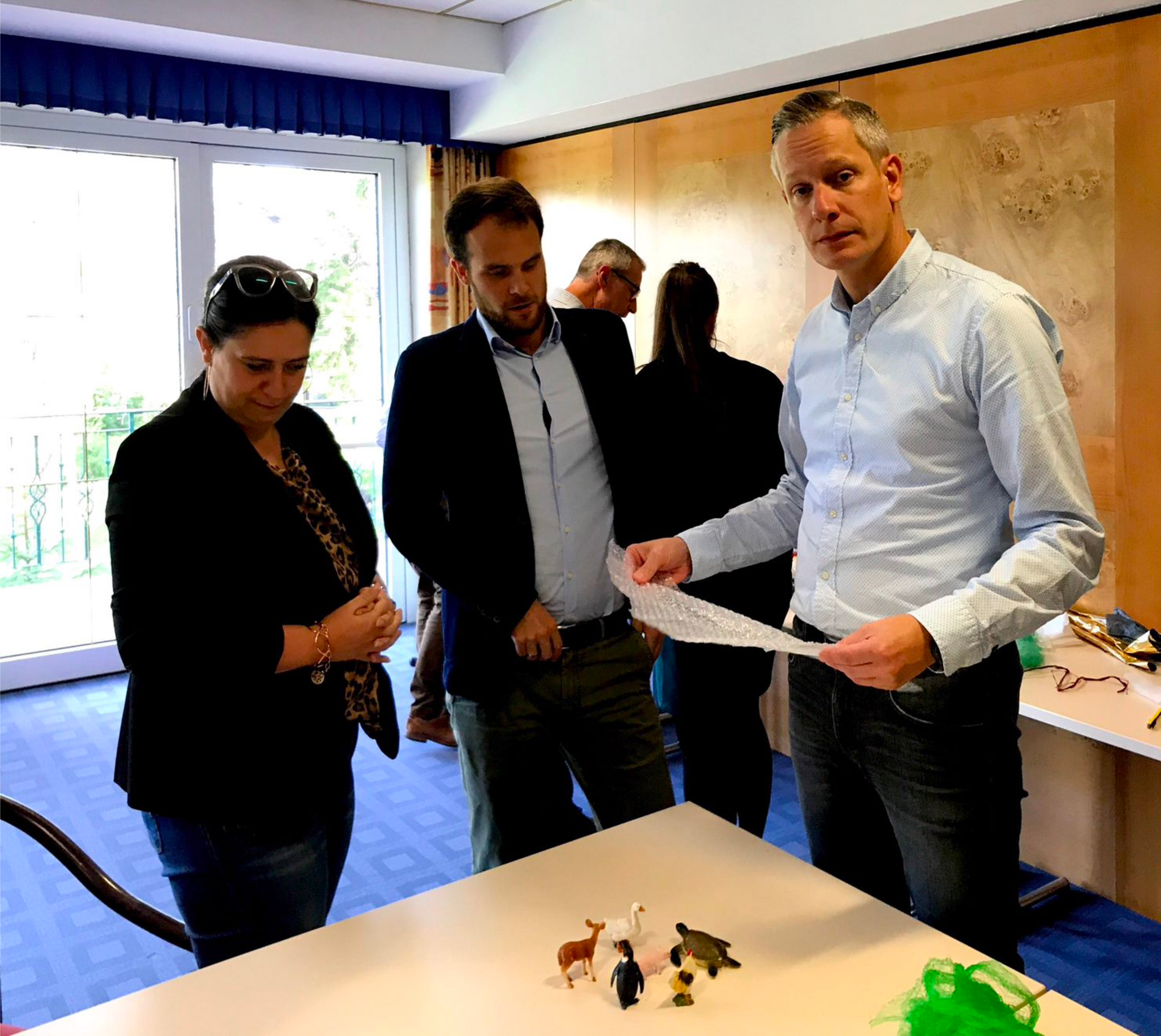 Drei Personen stehen an einem Tisch mit kleinen Tierfiguren, während ein Mann Luftpolsterfolie hält.
