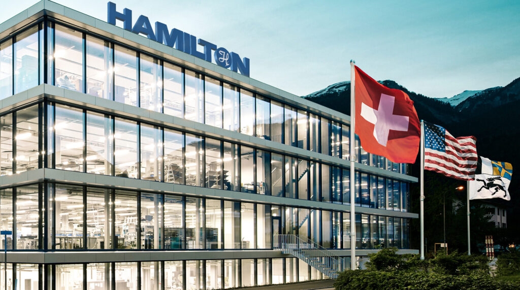 Modernes Hamilton Firmengebäude aus Glas mit wehenden Flaggen der Schweiz und der USA.