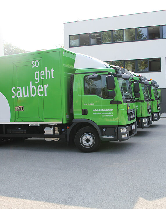 Grüne LKW der Firma Hollu stehen nebeneinander vor einem weißen Bürogebäude.