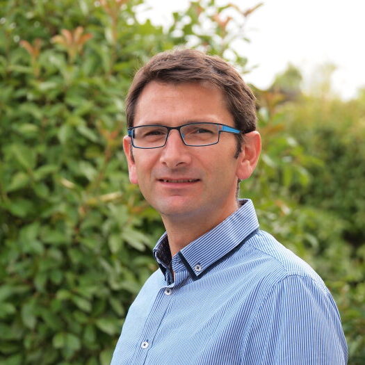 Ein lächelnder Mann mit Brille und blau gestreiftem Hemd vor grünem Hintergrund.