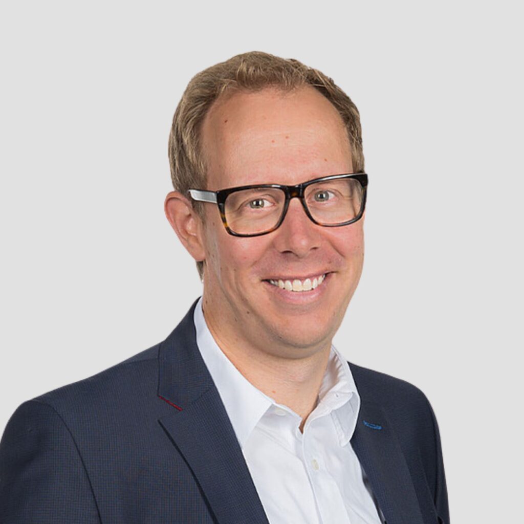 Ein lächelnder Mann mit Brille, weißem Hemd und dunkelblauem Sakko.