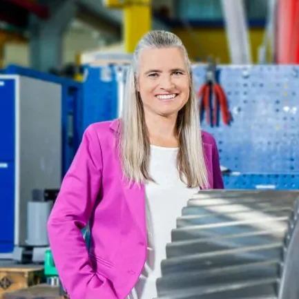 Lächelnde Frau im pinken Blazer steht neben einem großen Zahnrad in einer Werkstatt.