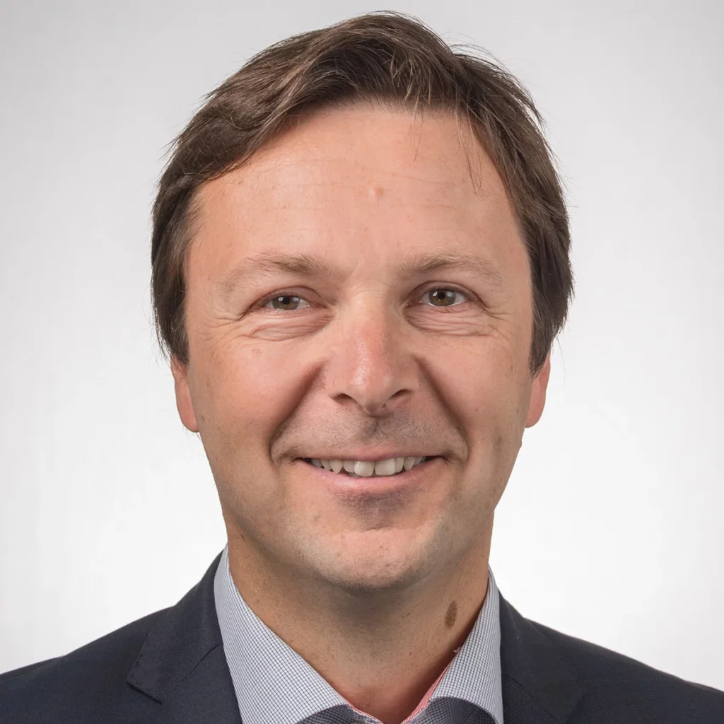 Ein freundlich lächelnder Mann im dunklen Anzug vor hellem Hintergrund.